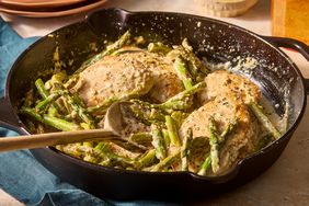One Pan Lemon Chicken and Asparagus in a cast iron skillet with a wooden spoon, with lemon wedges in the background 