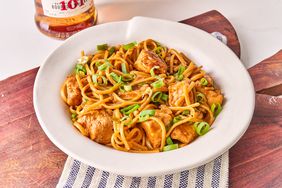 Bowl of One Pan Tipsy Chicken and Noodle garnished with green onions on a wooden surface, bottle of whiskey in the background