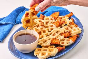 A plate of waffles and chicken with a bowl of syrup, a hand dipping a waffle segment