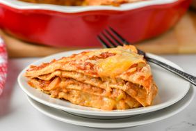 Layered casserole slice on a plate with cheese melted on top in front of a red dish