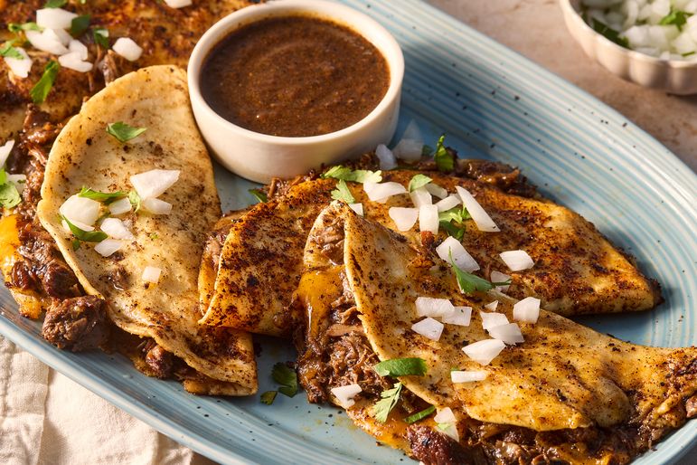 Quesabirria tacos on blue platter garnished with onions and cilantro next to a bowl with sauce 