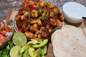 plate of shrimp and bell pepper fajitas with tortillas, sliced avocado, pico de gallo, and cream sauce on a board