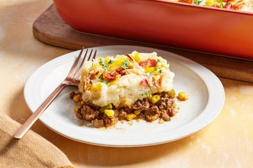 Slice of Cowboy Mashed Potato Casserole served on a white plate with more of the casserole in a red baking dish on a wooden board in the background 