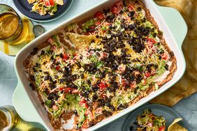 Baking dish with seven-layer dip topped with cheese, olives, and tomatoes.