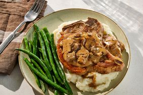 A pork chop topped with mushrooms and gravy, served over mashed potatoes with green beans